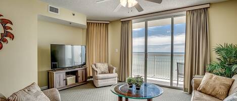 A living room with a flat-screen TV, two armchairs, a glass coffee table, and a ceiling fan.