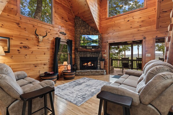 A cozy cabin interior with a stone fireplace, wooden walls, and a large window overlooking a forest.