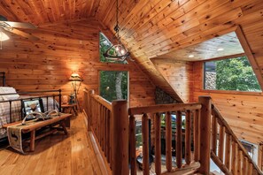 A wooden cabin interior with a loft bedroom, a fireplace, and a view of the outdoors.