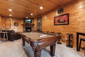A pool table in a room with wooden walls, a stone fireplace, and a television mounted above it.