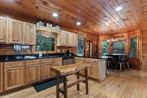A wooden cabin interior with a kitchen area featuring wooden cabinets, a central island, and a dining table with chairs. - Drink Up The View - Morganton (Morganton)