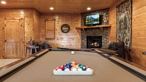 Ein Billardtisch mit Kugeln, ein darüber befestigter Fernseher, ein Steinkamin und eine Holzwand mit Spiegel.