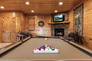 A pool table with balls, a television mounted above it, a stone fireplace, and a wooden wall with a mirror. - Drink Up The View - Morganton (Morganton)