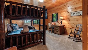 Ein Interieur einer Holzhütte mit Etagenbett, Schreibtisch, Schaukelstuhl und einem Fenster mit Blick auf Bäume.