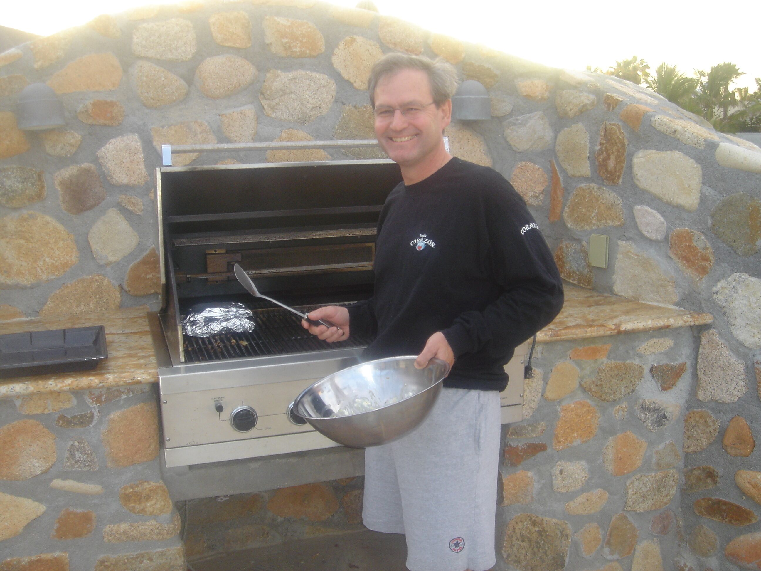 A person standing next to a stone-built outdoor grill with a metal bowl and spatula.
