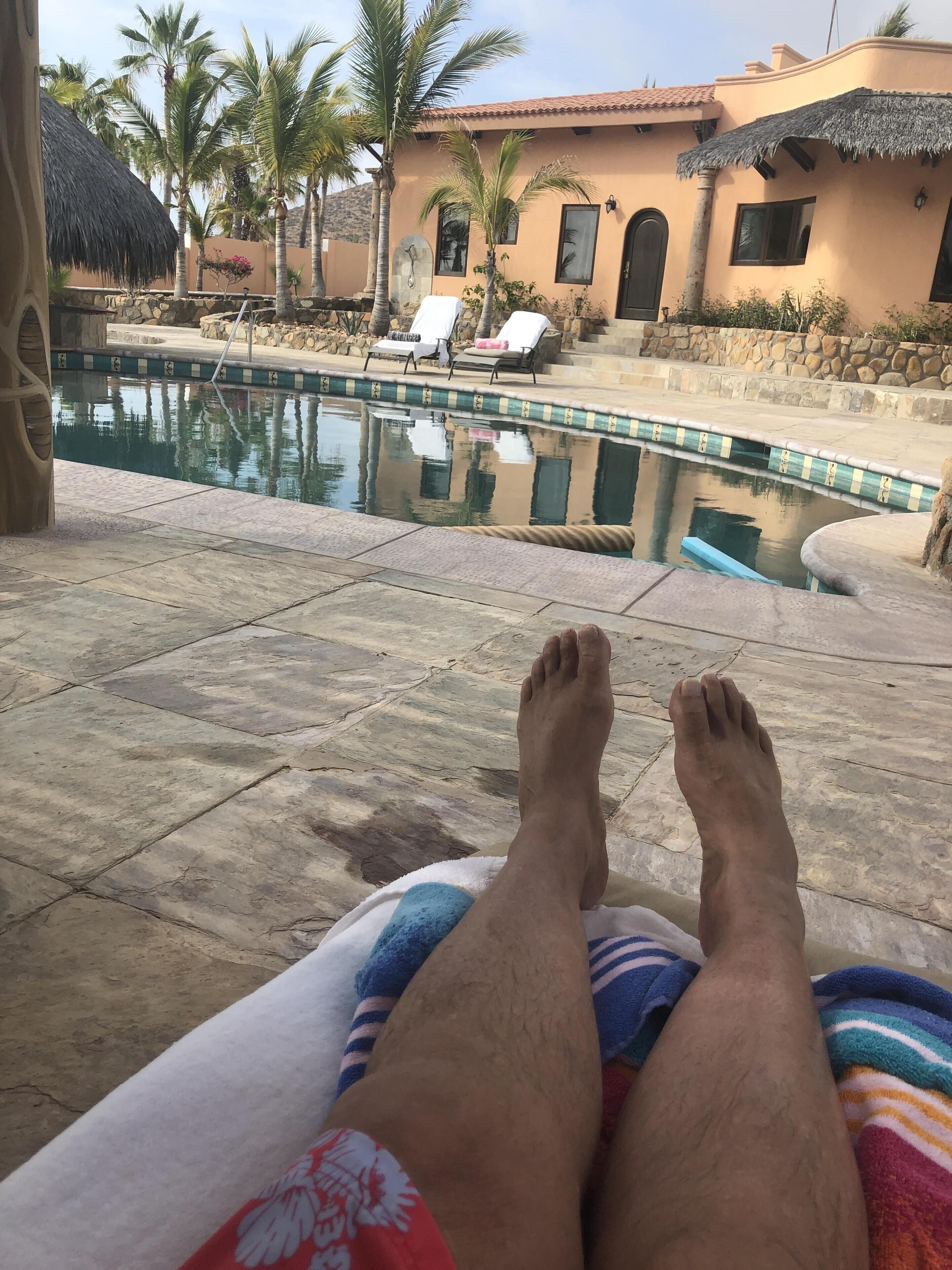 A poolside view with a lounge chair, a stone pool, and a building with a tiled roof.