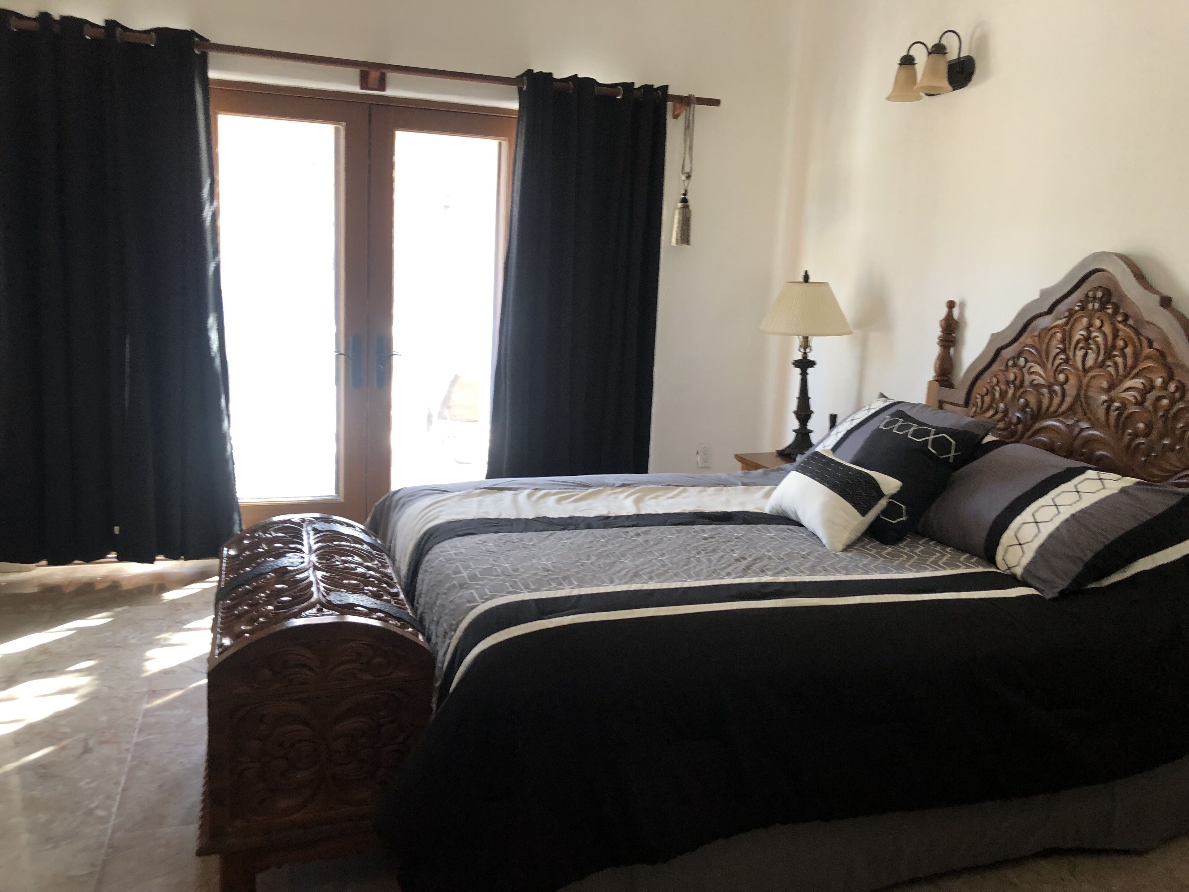 Quarto com uma cama grande, cabeceira de madeira, abajur e vista para o exterior através de uma porta.