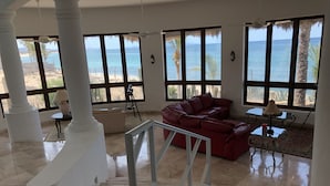 A spacious living area with a red leather sofa, a glass coffee table, and a view of the ocean through large windows. - McGann Estate Resort: Two Houses on the Beach, sleeps 14 (Los Barriles)