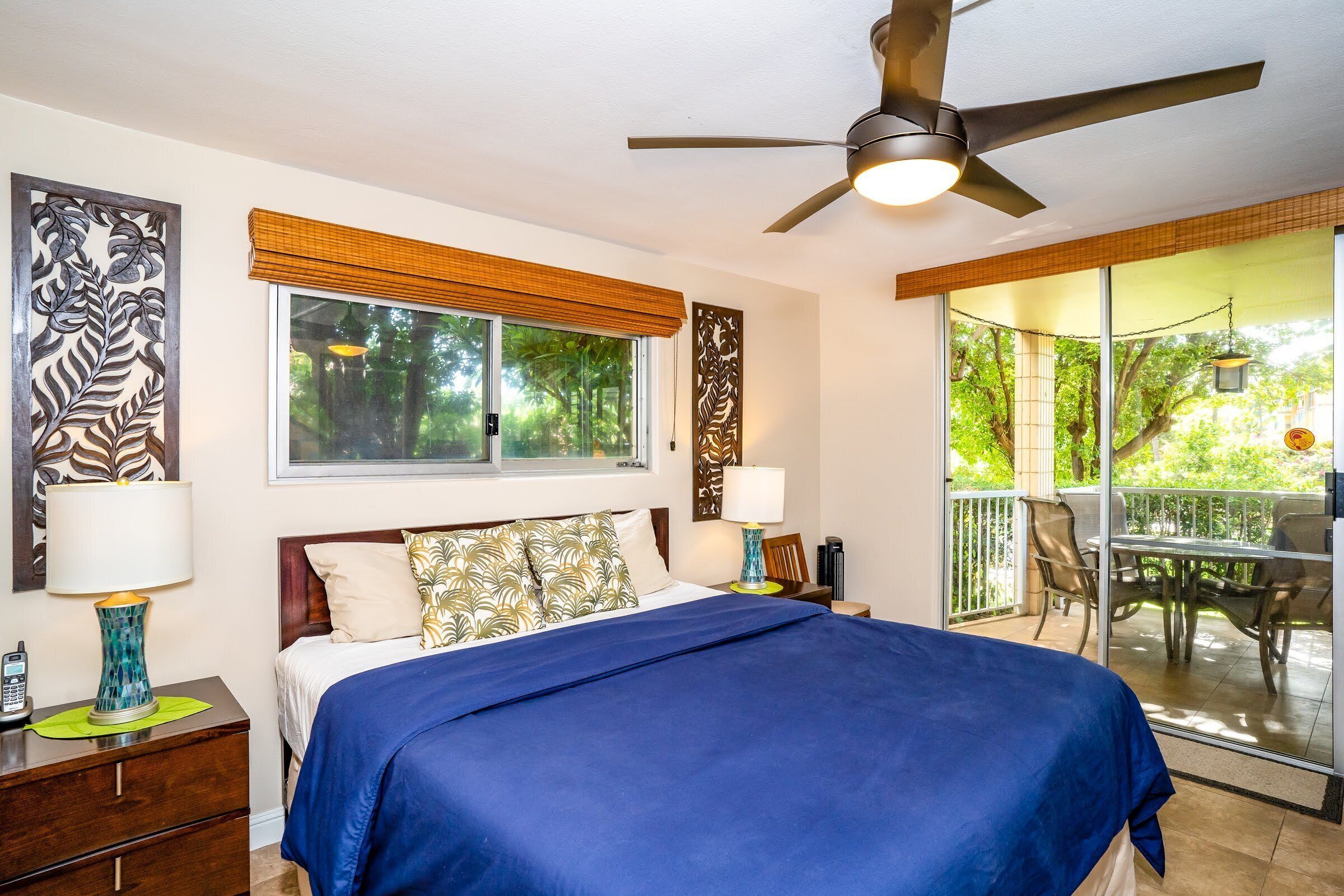A bedroom with a bed, a ceiling fan, a window with blinds, a nightstand, and a view of the outdoors.