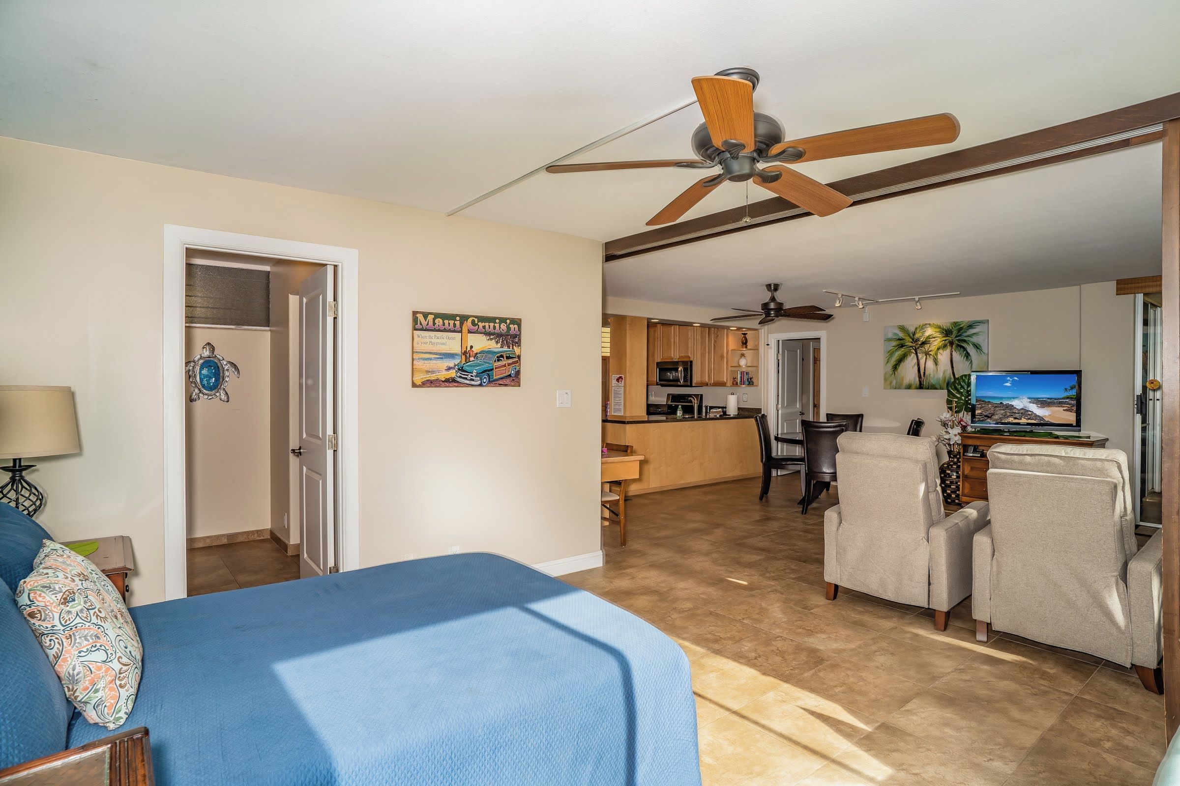 A spacious living area with a ceiling fan, a kitchen with wooden cabinets, a dining area with a TV, and a bedroom with a bed and a nightstand.