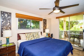 Ein Schlafzimmer mit Bett, Deckenventilator, Fenster mit Jalousien, Nachttisch und Blick ins Freie.