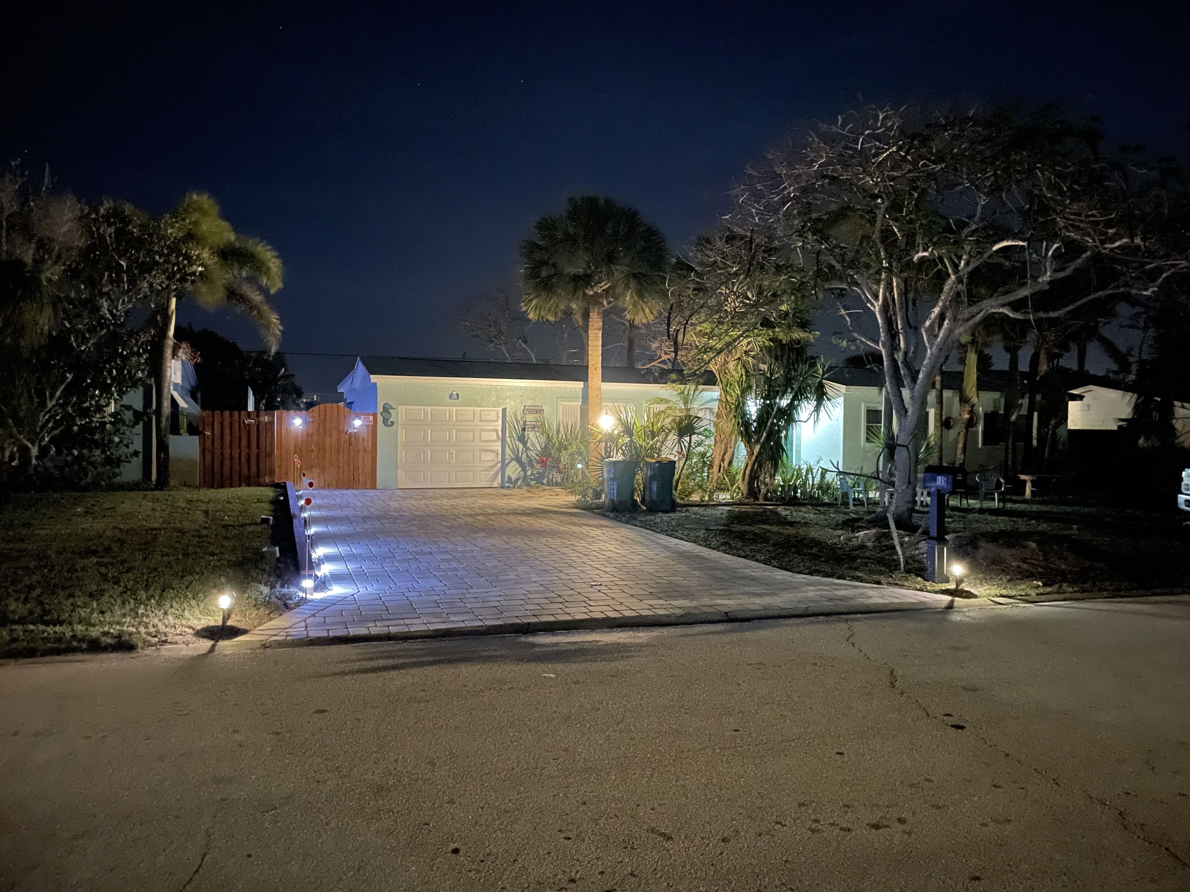 A driveway leading to a garage with a wooden fence on the left and a well-lit house entrance.