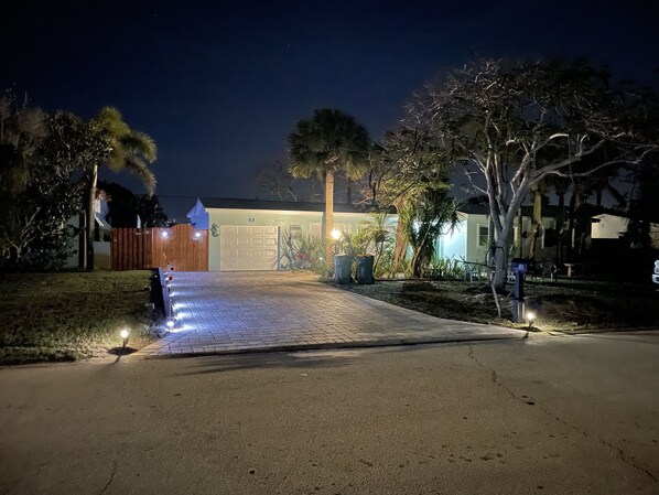 A driveway leading to a garage with a wooden fence on the left and a well-lit house entrance.