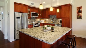 A modern kitchen with wooden cabinets, stainless steel appliances, a granite island, pendant lighting, and a painting on the wall. - Pili Mai 3B: Luxury Condo w/hot tub & pool, near beaches, golf & dining! (Koloa)