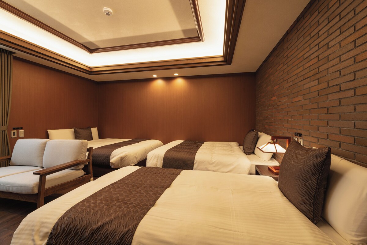 Habitación de hotel con tres camas, cabecera de madera, pared de ladrillo e iluminación empotrada en el techo.