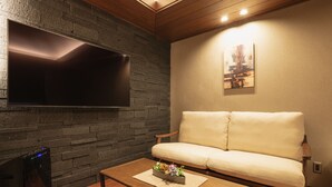 A living room with a flat-screen TV mounted on a brick wall, a beige sofa, and a wooden coffee table with a decorative centerpiece. - GRAND BASE Beppu (Beppu)