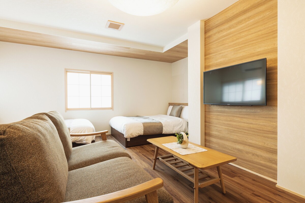 Una habitación moderna y bien iluminada, con suelo de madera, un sofá, una cama, un televisor montado en la pared y una ventana que proporciona luz natural.