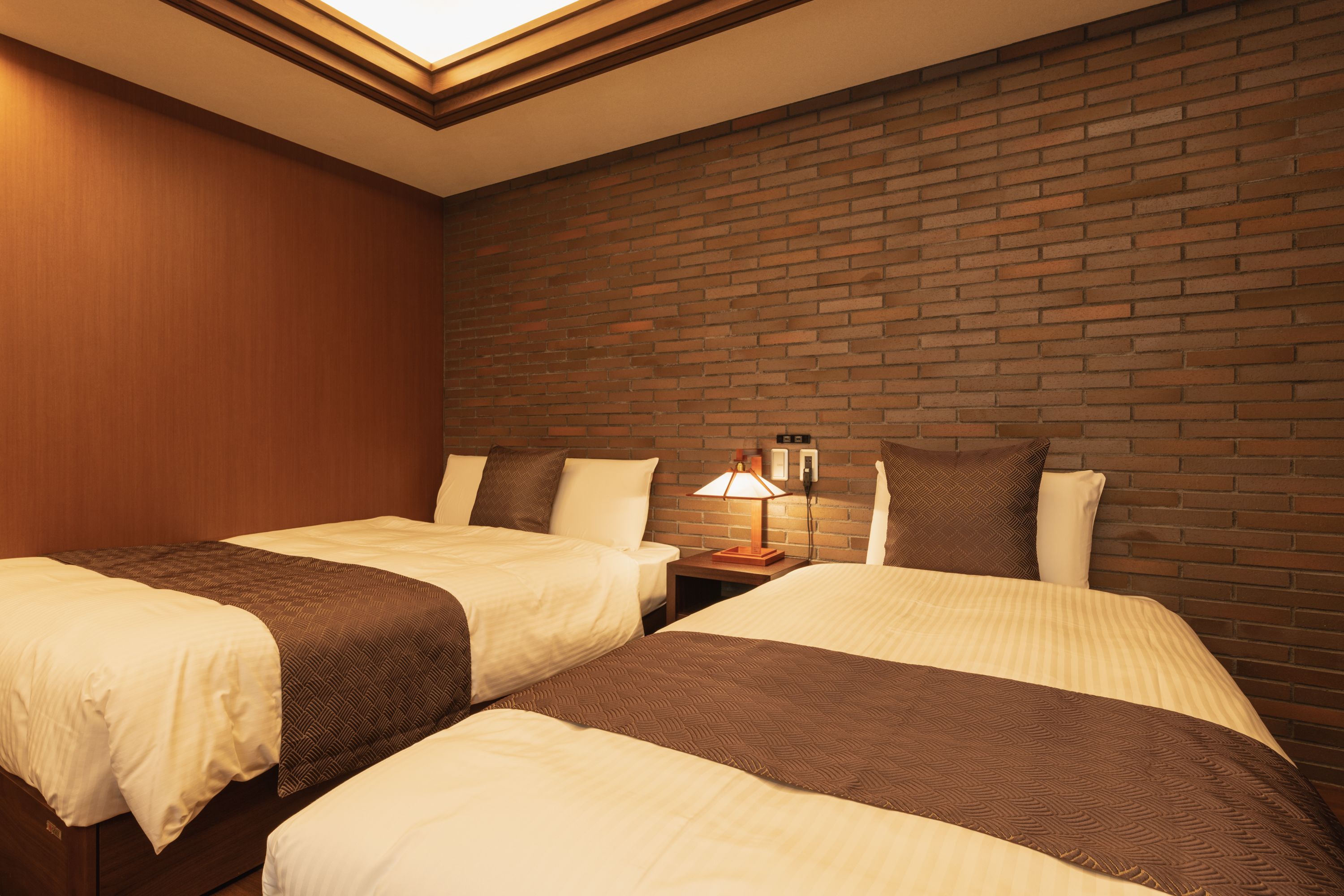 A hotel room with two beds, a brick wall, and a nightstand with a lamp.