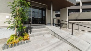 A modern building entrance with a glass door, a staircase, and a small garden area. - GRAND BASE Beppu (Beppu)