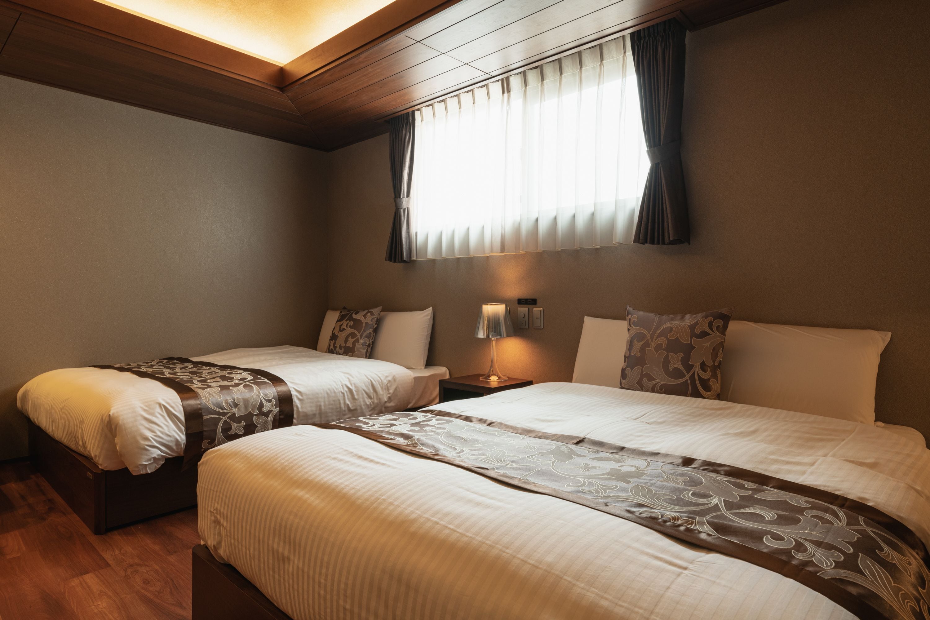 A hotel room with two beds, wooden ceiling, and a window with curtains.