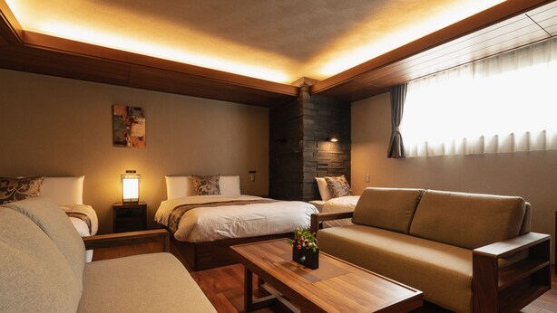 A modern hotel room with a wooden ceiling, a large bed, a sofa, a small table, and a window with curtains. - GRAND BASE Beppu (Beppu)