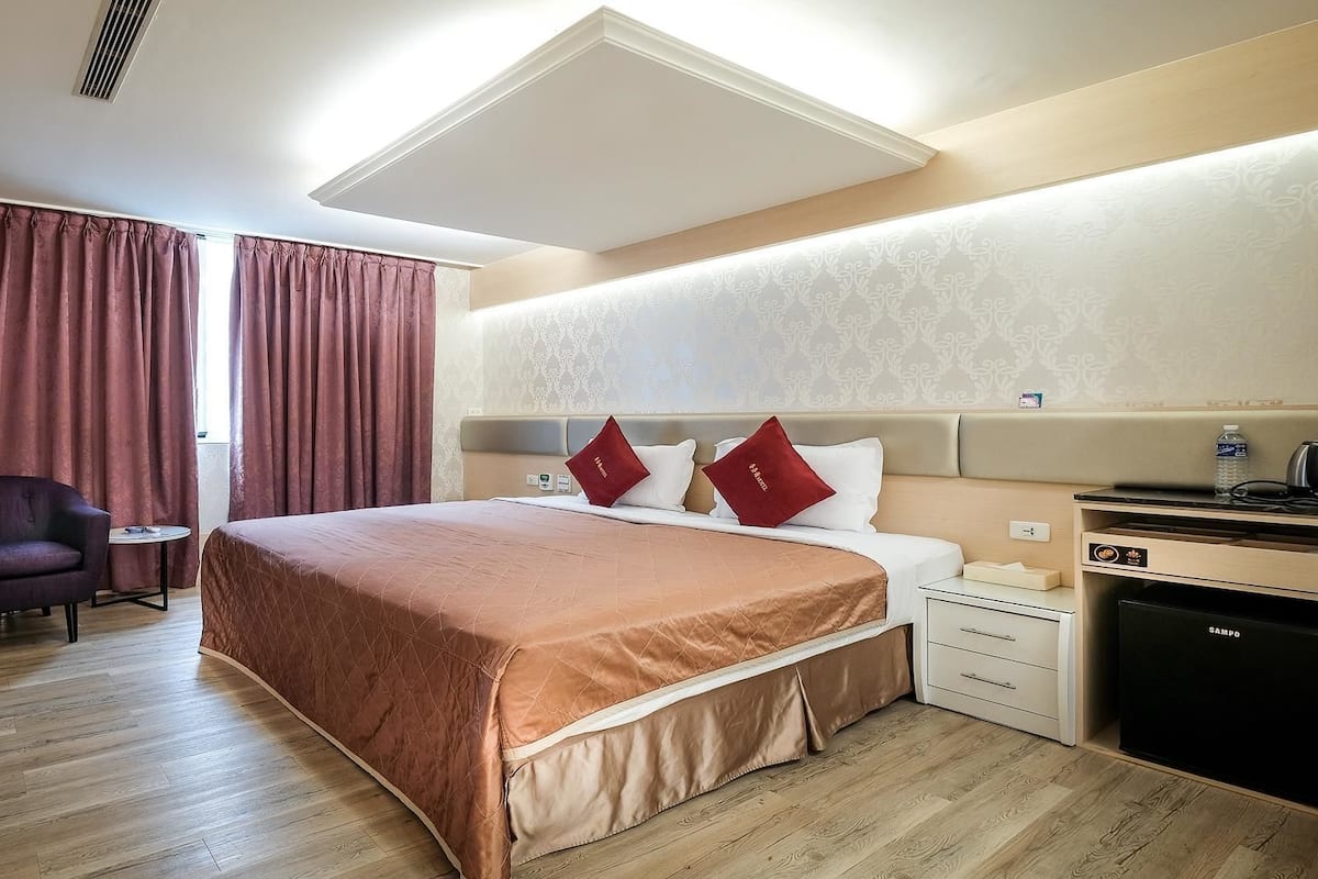 A hotel room with a bed, a chair, a small table, a desk, and a minibar.