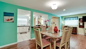 A dining area with wicker chairs and a glass table, a kitchen with white cabinets, and a living area with a television and artwork.