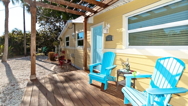 A wooden deck with blue Adirondack chairs, a small table with drinks, and a house with a blue door and window blinds.