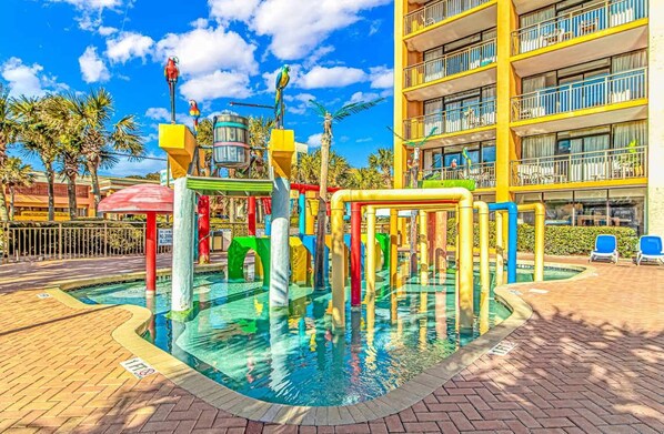 A colorful play area with a pool, palm trees, and a multi-story building in the background.