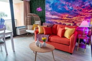 A living room with a red sofa, a wicker chair, a round wooden table with vases, and a wall mural.