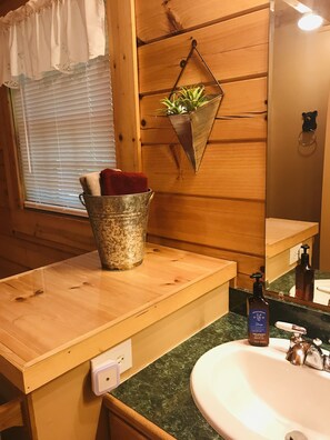 A wooden cabin bathroom with a wooden vanity, a sink, a mirror, a potted plant, and a metal bucket with towels.