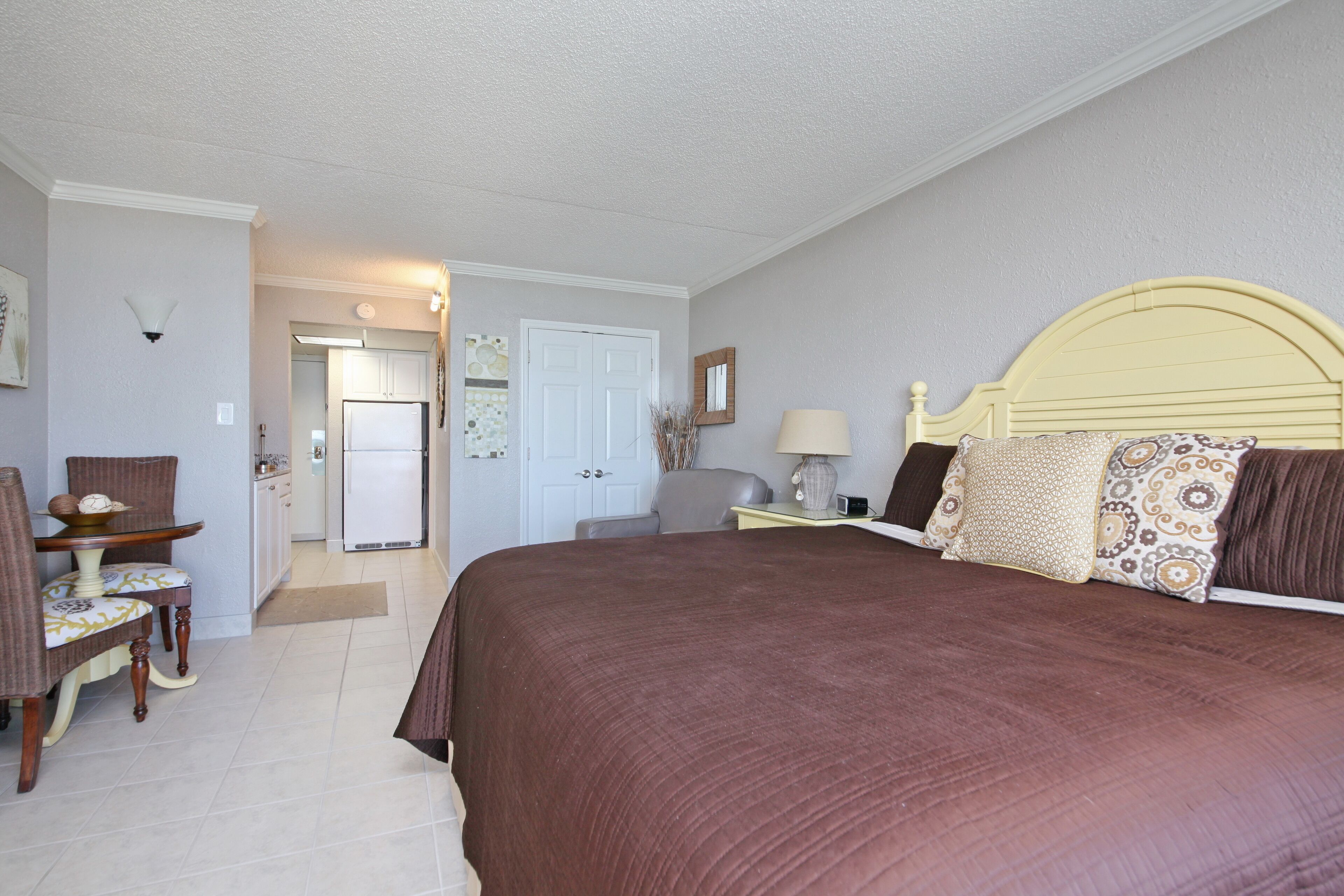 A bedroom with a large bed, a desk with a chair, a lamp, and a door leading to another room.