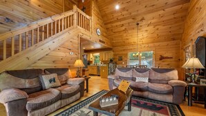 Interior acolhedor de uma cabana, com duas sofás, uma escada de madeira e um espaço de refeições.