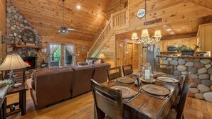 Interior acolhedor de cabana, com lareira de pedra, mesa de jantar de madeira e escada que dá para um mezanino.