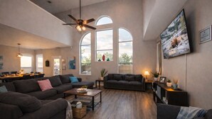 Ein geräumiges Wohnzimmer mit einer großen Sofagarnitur, einem Couchtisch und einem an der Wand montierten Fernseher. Große Fenster sorgen für natürliches Licht und bieten einen Blick ins Freie.