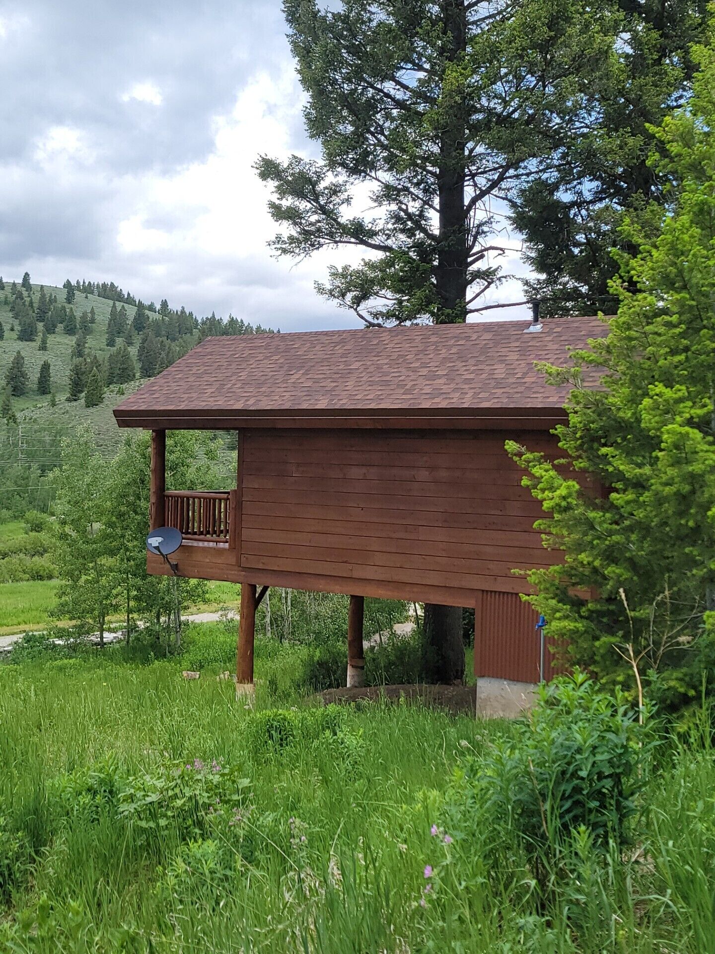 A wooden cabin with a balcony, surrounded by lush greenery and a hilly landscape.