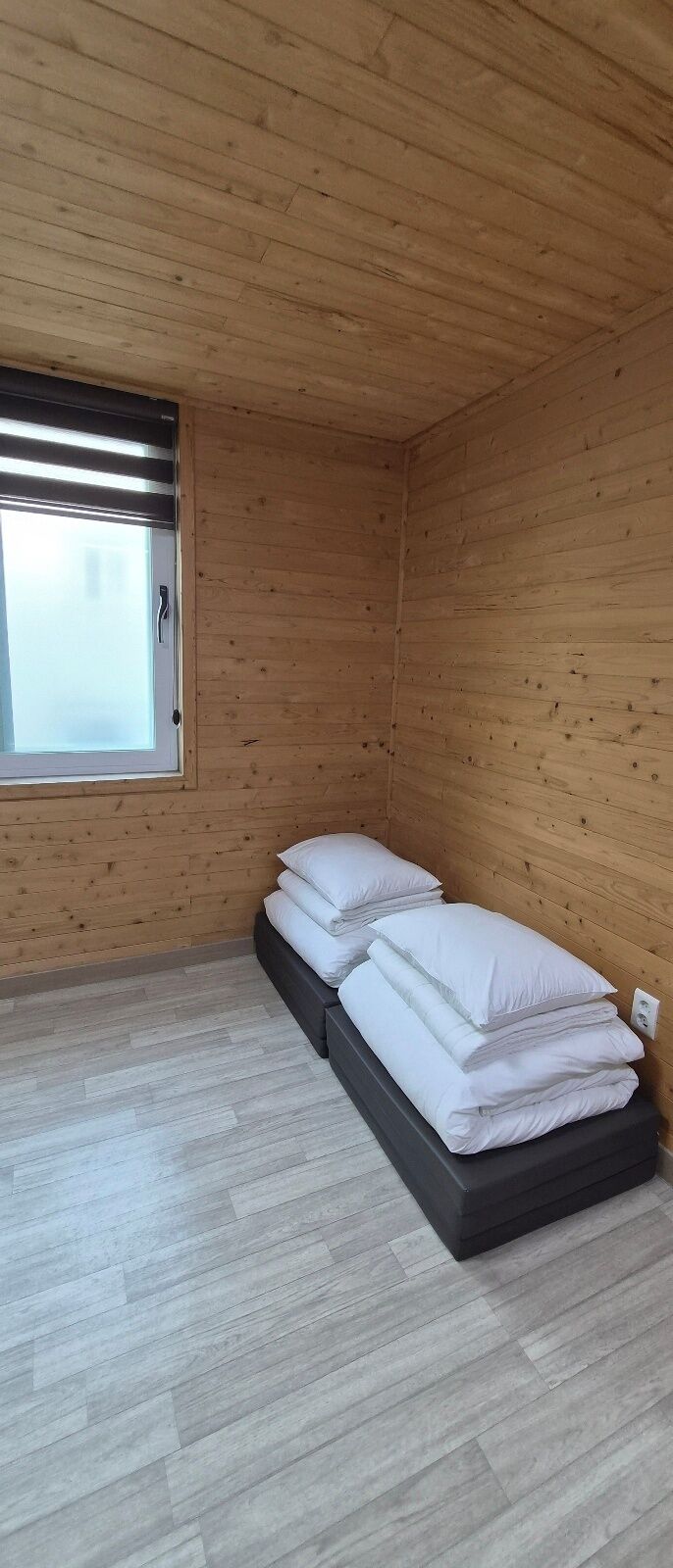 Een kamer met houten wanden, een bed met witte lakens, een raam met zonwering en een lichtschakelaar aan de muur.