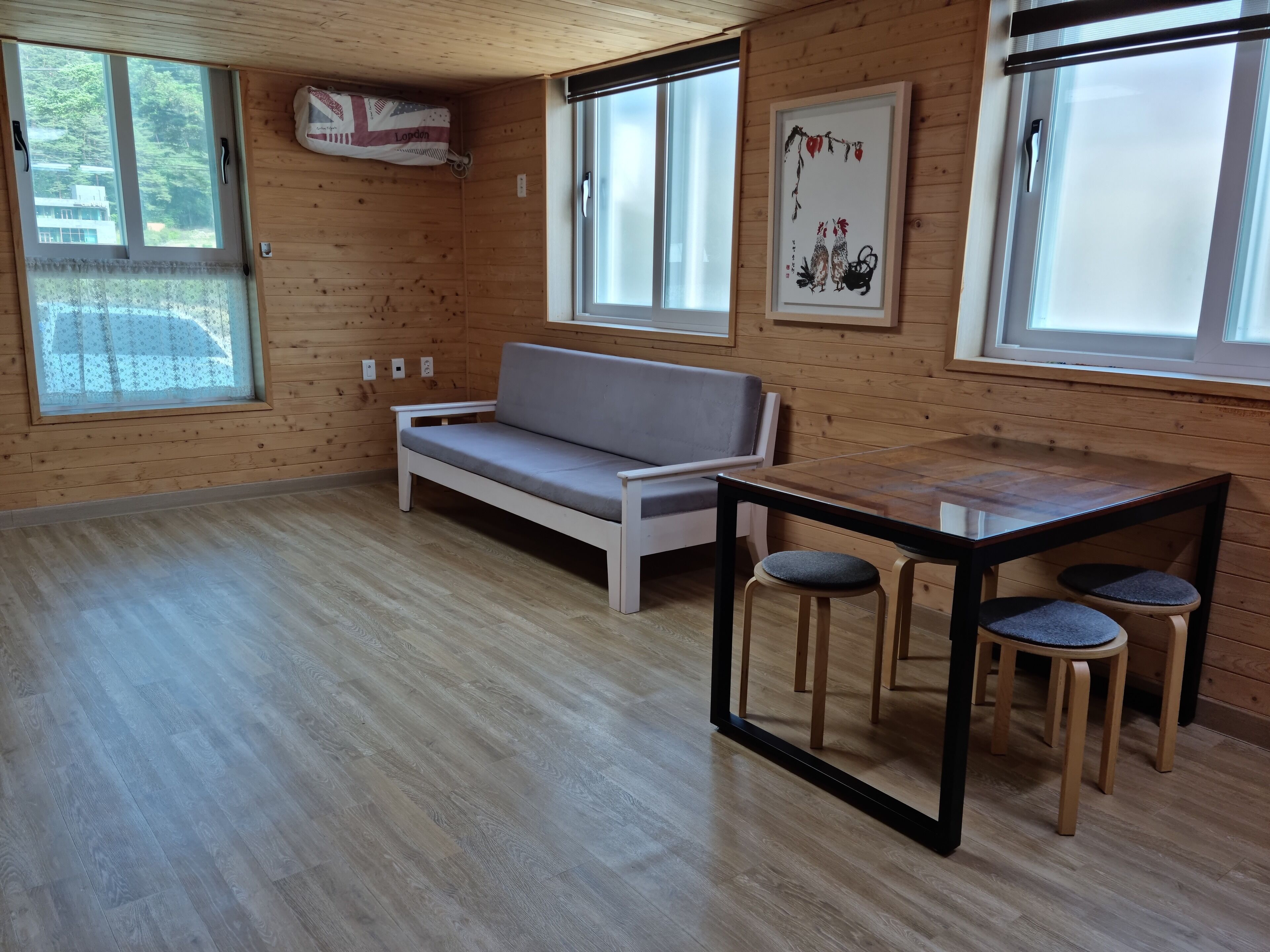 Una habitación con paredes y suelos de madera, un sofá blanco, una mesa de vidrio con taburetes y ventanas amplias.