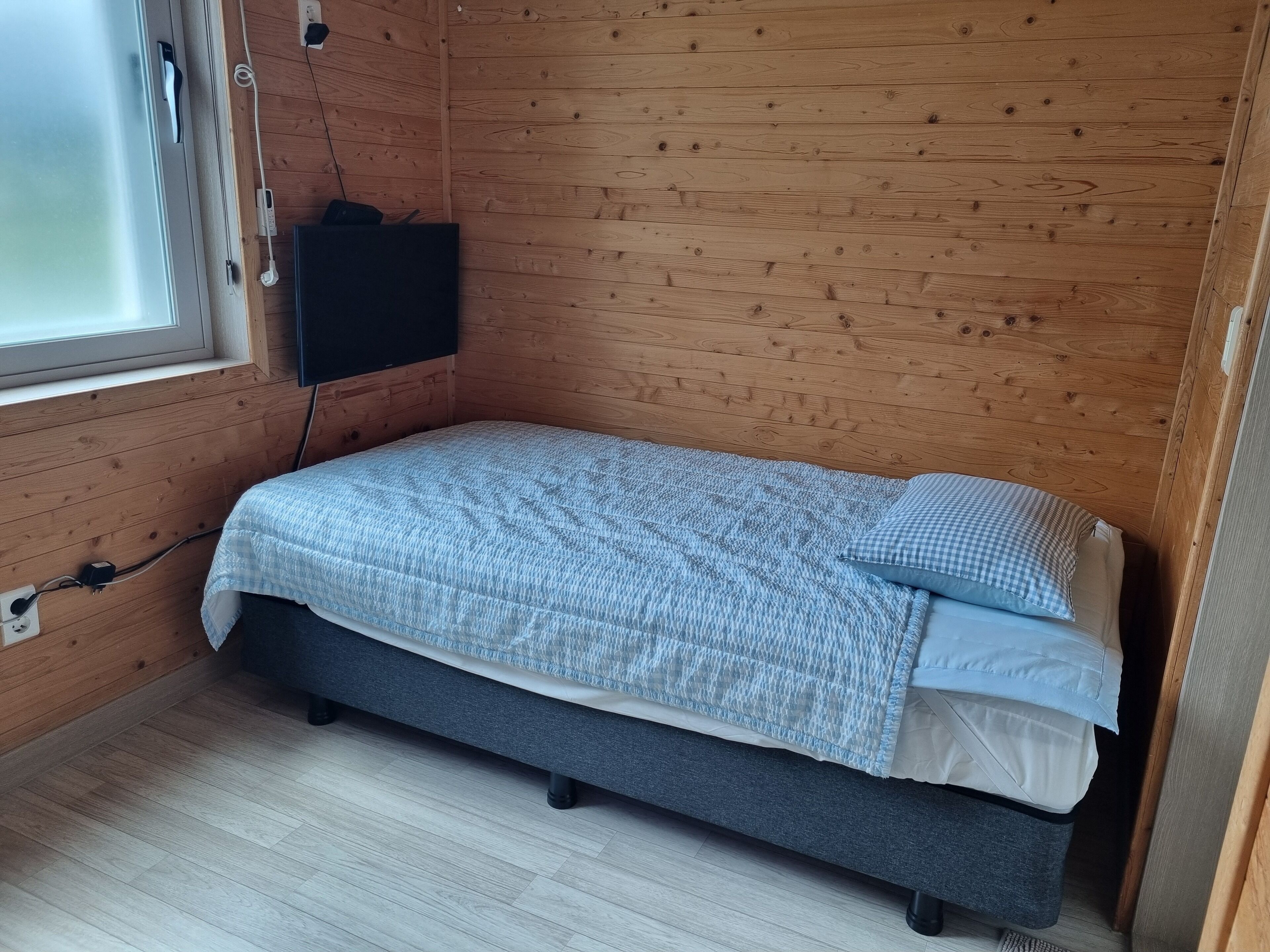 A small, single-bed room with a wooden interior, a window, and a power outlet on the wall.