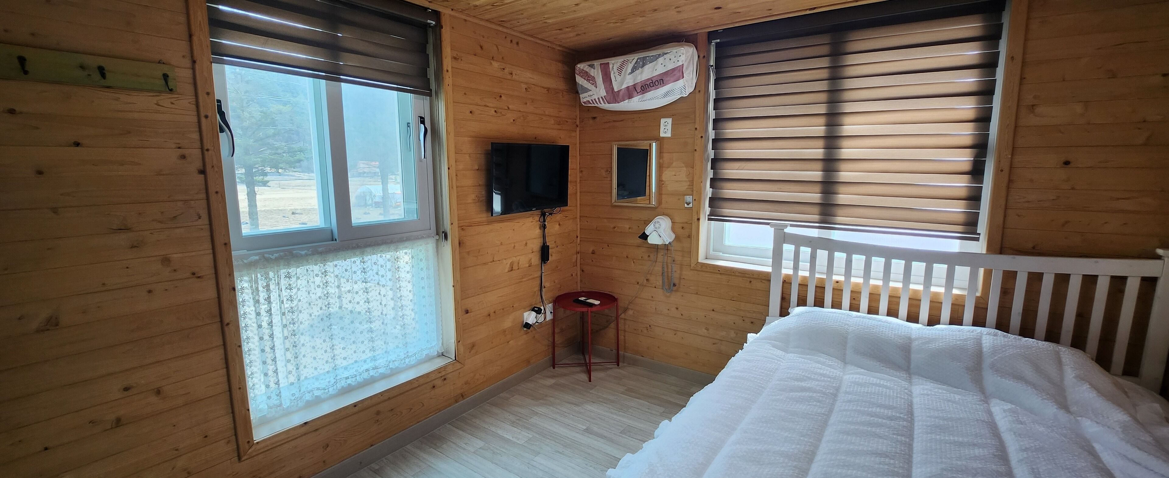 A wooden cabin room with a bed, a TV, a telephone, and a window with a curtain.
