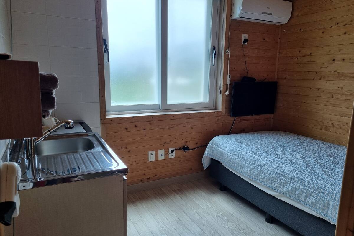 Een compacte kamer met houten inrichting, een klein keukentje met een spoelbak en kookplaat, een enkel bed en een raam met gesatineerd glas.