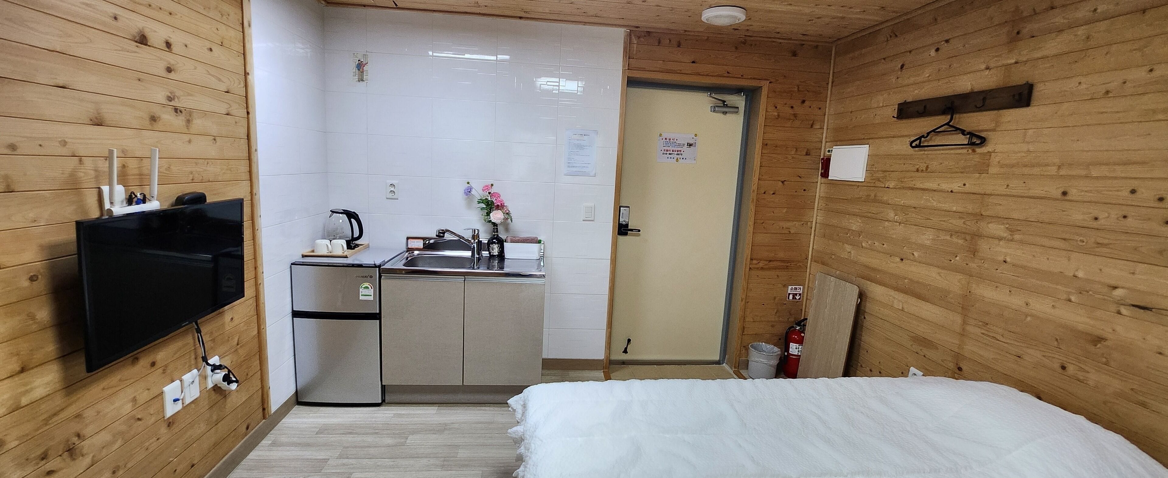 A compact room with a bed, a TV mounted on the wall, a small kitchenette, and a wooden door.