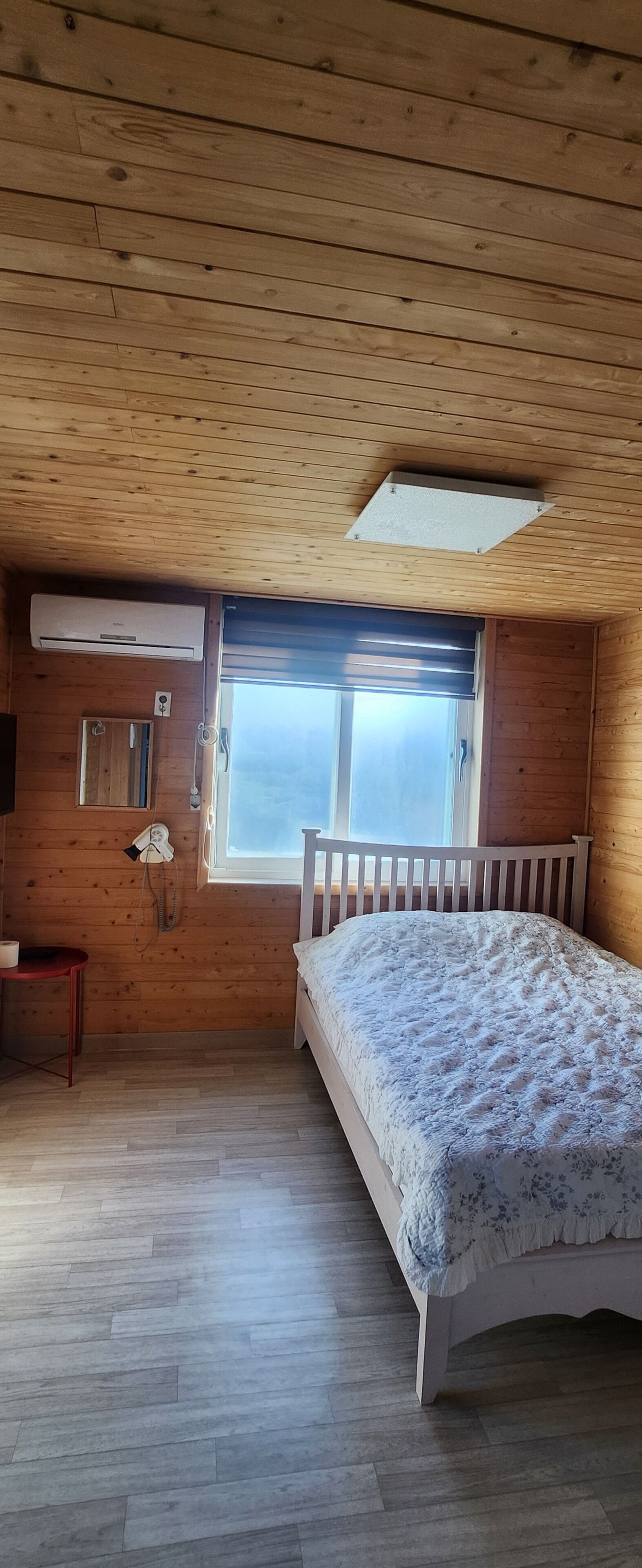 A bedroom with a wooden ceiling, a window with blinds, a bed with a floral bedspread, a bedside table, and a wall-mounted heater.