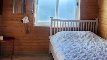 A bedroom with a wooden ceiling, a window with blinds, a bed with a floral bedspread, a bedside table, and a wall-mounted heater.