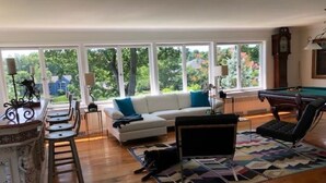 A living room with a white sofa, a pool table, a dog, and large windows offering a view of trees and a house.