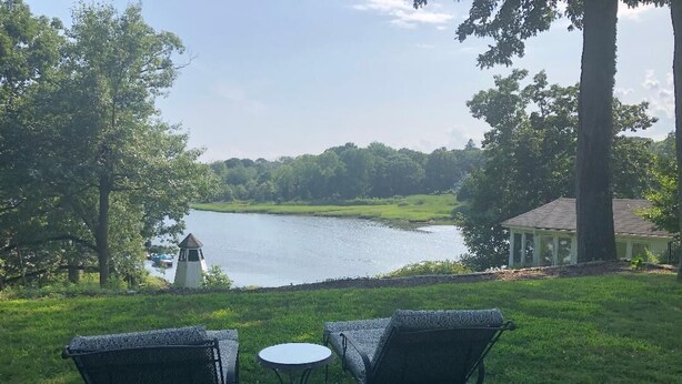 A grassy area with two metal chairs and a small round table, overlooking a lake with a gazebo in the background.
