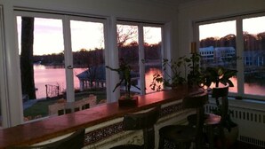 A room with large windows overlooking a body of water, a bar area with stools, and potted plants.
