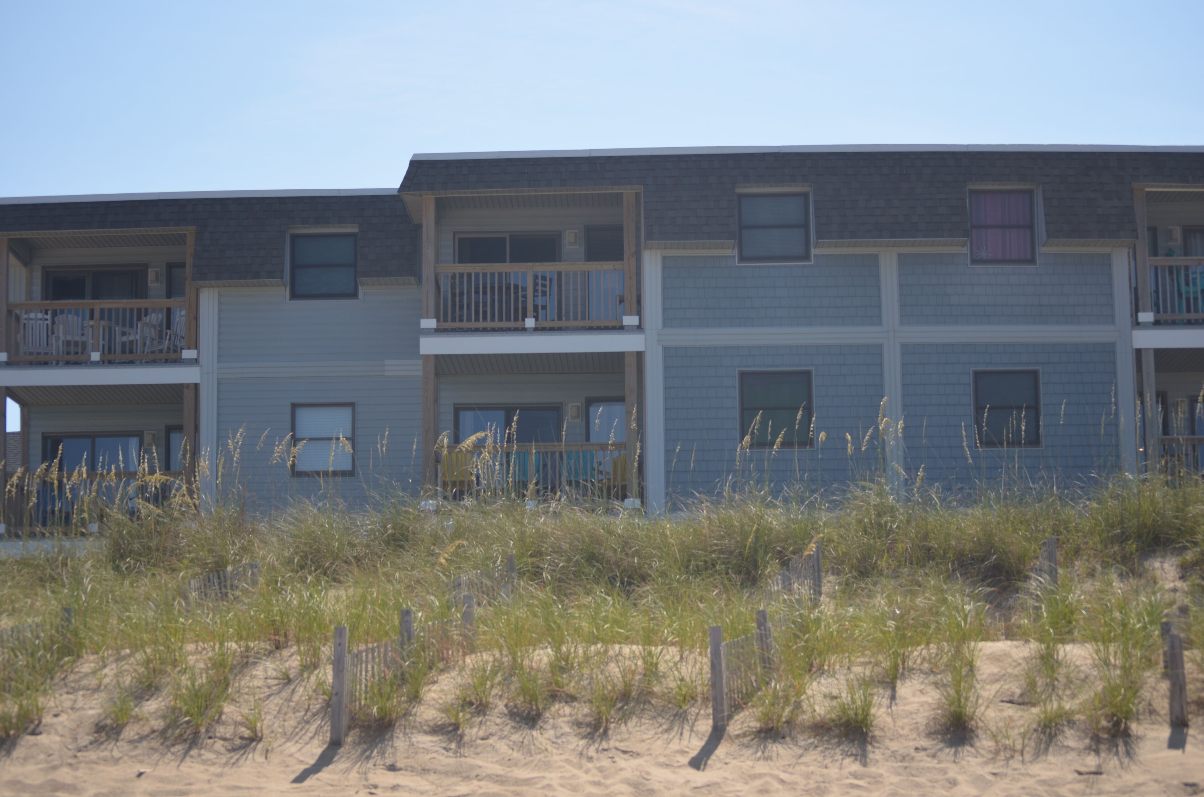A multi-story building with balconies, located near a sandy area with vegetation.