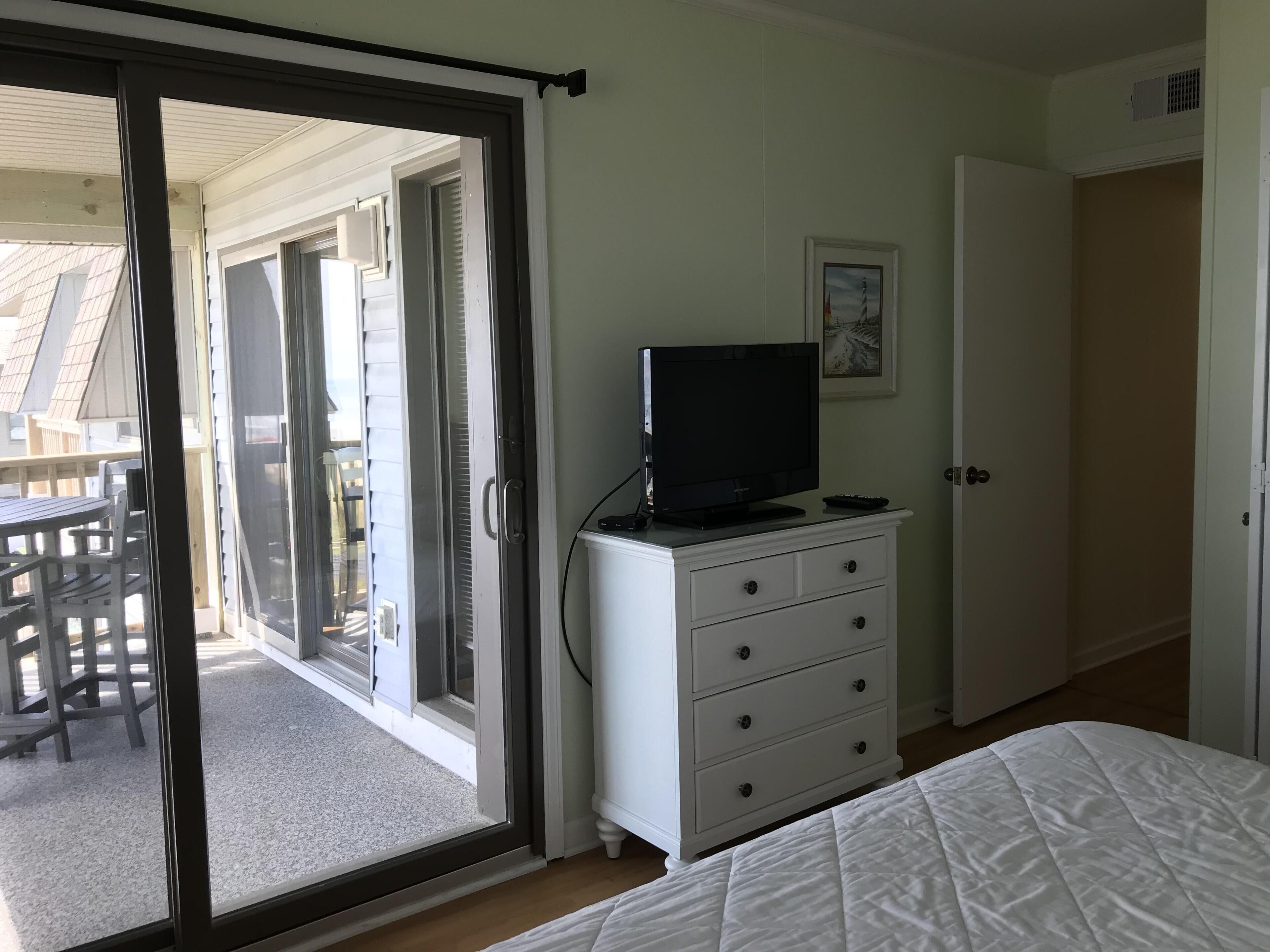 Une chambre avec une porte-fenêtre coulissante donnant sur un balcon, une commode blanche, un lit et un tableau encadré au mur.