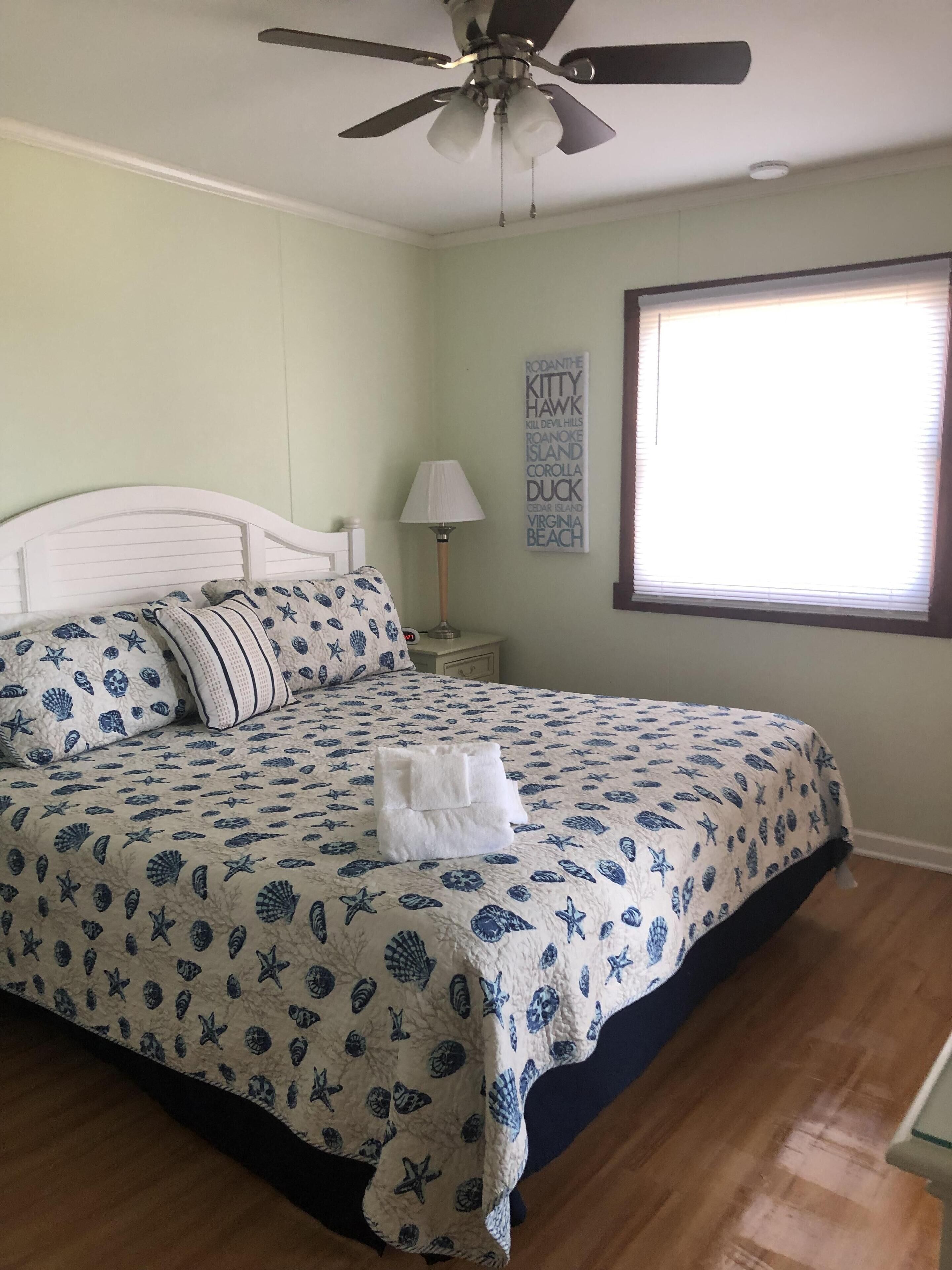 Un dormitorio con cama, ventilador de techo, mesita de noche con lámpara, ventana con persianas y pared con una nube de palabras.